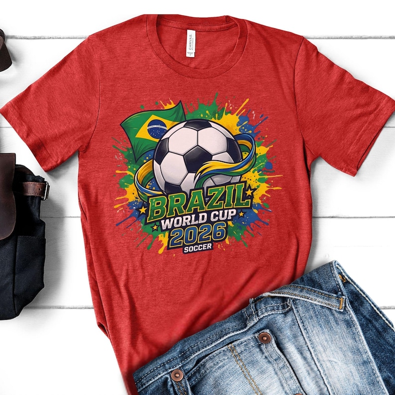 K&ouml;nnte beinhalten: Rotes T-Shirt mit einem Fu&szlig;ball-Grafik, der brasilianischen Flagge und dem Text "BRAZIL WORLD CUP 2026 SOCCER". Das Design umfasst farbenfrohe Farbspritzer in Gelb, Blau und Gr&uuml;n. Das Shirt wird mit einer blauen Jeans kombiniert.