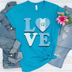 May include: A turquoise t-shirt with the word "LOVE" in light blue letters. The "O" is a heart with the Guatemalan flag design. The shirt is paired with a denim jacket and jeans.