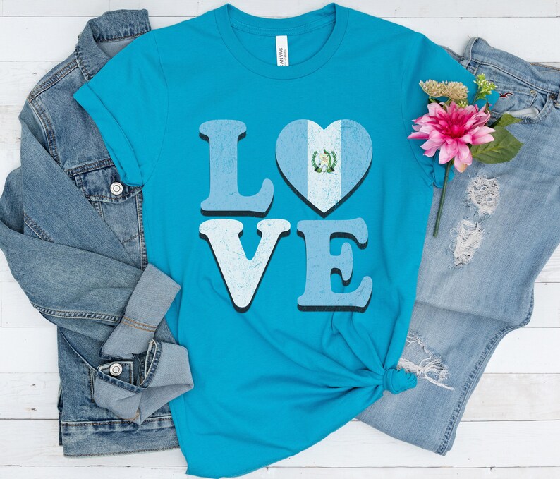 May include: A turquoise t-shirt with the word "LOVE" in light blue letters. The "O" is a heart with the Guatemalan flag design. The shirt is paired with a denim jacket and jeans.