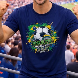K&ouml;nnte beinhalten: Marineblaues T-Shirt mit einem Fu&szlig;ball-Design und dem Text "BRAZIL WORLD CUP 2026 SOCCER". Das Design beinhaltet die brasilianische Flagge und farbenfrohe Farbspritzer. Eine Person tr&auml;gt das T-Shirt.