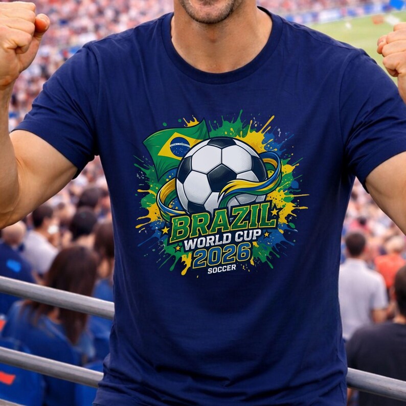 K&ouml;nnte beinhalten: Marineblaues T-Shirt mit einem Fu&szlig;ball-Design und dem Text "BRAZIL WORLD CUP 2026 SOCCER". Das Design beinhaltet die brasilianische Flagge und farbenfrohe Farbspritzer. Eine Person tr&auml;gt das T-Shirt.