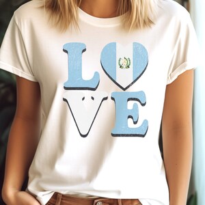 May include: Cream-coloured t-shirt featuring the word "LOVE" in a distressed font. The letters "L" and "E" are light blue, the "V" is white, and the "O" is a heart shape with the Guatemalan flag design. The shirt has a vintage appearance.