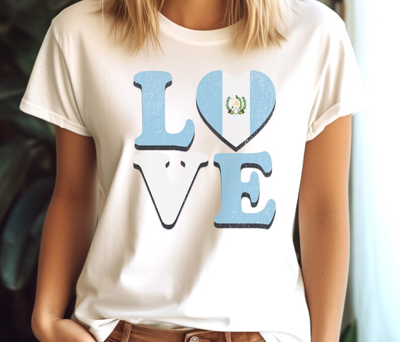 May include: Cream-coloured t-shirt featuring the word "LOVE" in a distressed font. The letters "L" and "E" are light blue, the "V" is white, and the "O" is a heart shape with the Guatemalan flag design. The shirt has a vintage appearance.