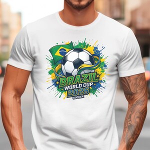 K&ouml;nnte beinhalten: Wei&szlig;es T-Shirt mit einem Fu&szlig;ball-Design, der brasilianischen Flagge und dem Text "BRAZIL WORLD CUP 2026 SOCCER". Das Design enth&auml;lt gr&uuml;ne, gelbe und blaue Akzente und ein stilisiertes Band um den Fu&szlig;ball.