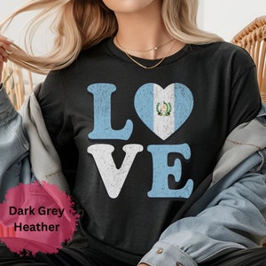 May include: Dark grey heather t-shirt featuring the word "LOVE" in white and light blue block letters. The "O" is a heart shape with the Guatemalan flag. The shirt has a distressed, vintage appearance.