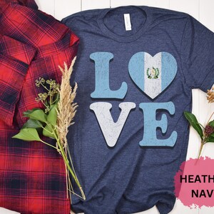 May include: Navy blue t-shirt featuring the word "LOVE" in light blue and white, with the "O" replaced by a heart displaying the Guatemalan flag. A red and black plaid shirt is draped on the left.