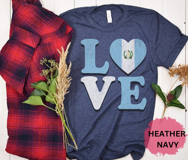 May include: Navy blue t-shirt featuring the word "LOVE" in light blue and white, with the "O" replaced by a heart displaying the Guatemalan flag. A red and black plaid shirt is draped on the left.