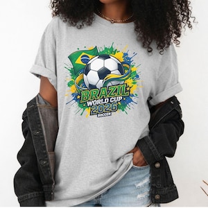 K&ouml;nnte beinhalten: Hellgraues T-Shirt mit einem Fu&szlig;ball-Design und dem Text "BRAZIL WORLD CUP 2026 SOCCER". Das Design beinhaltet die brasilianische Flagge und farbenfrohe Farbspritzer in Gr&uuml;n, Gelb und Blau.