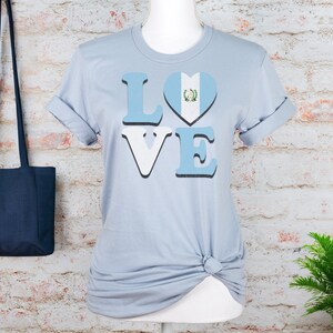 May include: Pale blue t-shirt featuring the word "LOVE" in a bold font, with the letter "O" replaced by a heart displaying the Guatemalan flag. The t-shirt is displayed on a mannequin against a brick wall.