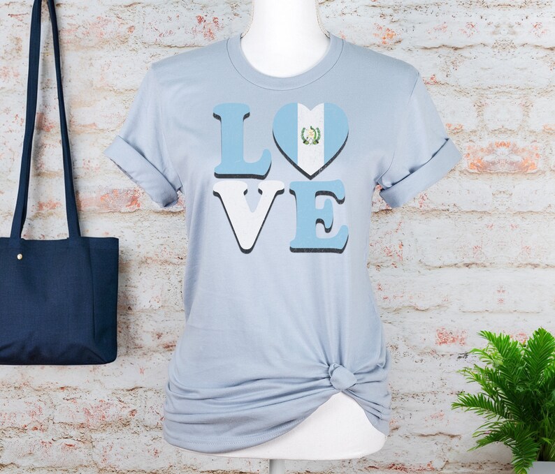 May include: Pale blue t-shirt featuring the word "LOVE" in a bold font, with the letter "O" replaced by a heart displaying the Guatemalan flag. The t-shirt is displayed on a mannequin against a brick wall.