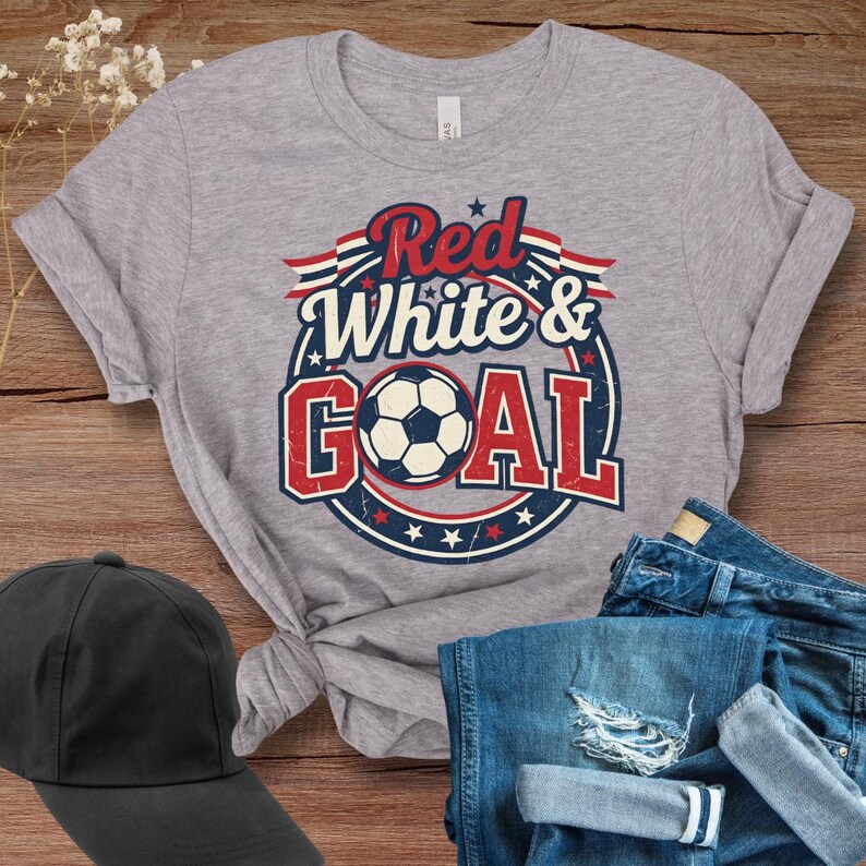 May include: A heather gray t-shirt with a graphic that says "Red, White & Goal" in red and blue, with a soccer ball in the center. The shirt is paired with blue jeans and a black baseball cap.