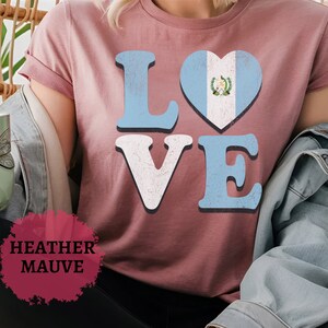 May include: Mauve t-shirt featuring the word "LOVE" in a distressed font. The "O" is a heart with the Guatemalan flag design. The shirt has short sleeves and is made of a soft material. The text "Heather Mauve" is at the bottom.