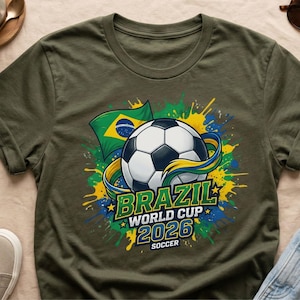 K&ouml;nnte beinhalten: Olivgr&uuml;nes T-Shirt mit einem Fu&szlig;ball, der brasilianischen Flagge und dem Text "BRAZIL WORLD CUP 2026 SOCCER". Das Design beinhaltet Farbspritzer in Gelb, Gr&uuml;n und Blau.