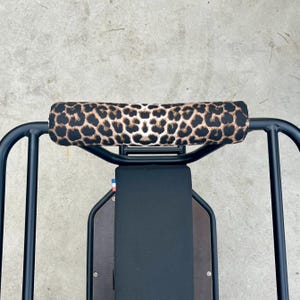 Può includere: Un portapacchi per bicicletta in metallo nero con un cuscino imbottito con stampa leopardata. Il cuscino è marrone e beige con un motivo maculato. Il portapacchi è montato su un sedile nero.