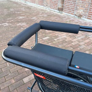 Può includere: Bicicletta cargo nera con una sella e uno schienale imbottiti. La bicicletta ha un telaio in metallo nero e un cestino in rete nera. La bicicletta ha un adesivo della bandiera francese sul retro.