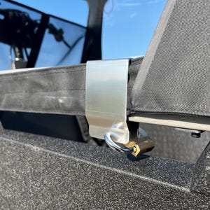 Peut inclure: Gros plan sur une pince métallique argentée fixant un panneau en tissu noir. Un petit cadenas doré est attaché à la pince, suggérant un mécanisme de verrouillage. L'arrière-plan montre un aperçu d'un ciel bleu.