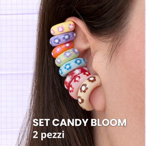 Può includere: Un set di polsini per orecchie colorati con motivi floreali. I polsini sono impilati sull'orecchio, mostrando una varietà di colori tra cui giallo, viola, arancione, verde, rosso e beige. Il testo "SET CANDY BLOOM" e "2 pezzi" è visibile.