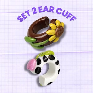 Può includere: Set di due orecchini a polsino. Uno è marrone con un girasole e viti verdi. L'altro è bianco con macchie nere e un tulipano rosa. Il testo "SET 2 EAR CUFF" è in viola.