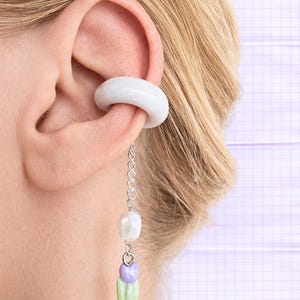 Può includere: Un set composto da un ear cuff bianco e un orecchino pendente. L'ear cuff è una fascia spessa e arrotondata. L'orecchino presenta una catena d'argento con una perla e un gruppo di perline viola e verdi. Il gioiello è indossato sull'orecchio di una persona.