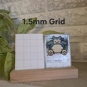 May include: A wooden display stand holding a grid and a Pokemon card featuring a cross-stitch design of Snorlax. The card is displayed against a white brick wall. Text on the card includes "Voraciousness" and "Thudding Press".