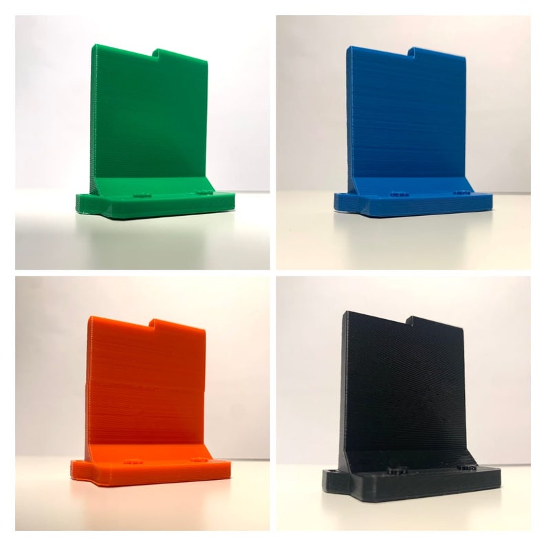 May include: Four 3D printed phone stands in different colours: green, blue, orange, and black. Each stand has a rectangular base and a vertical back.