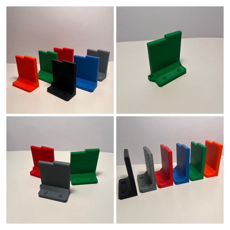May include: A set of six 3D printed phone stands in various colours, including red, black, blue, green, grey, and orange. The stands are designed to hold a phone upright.