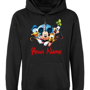 Puede incluir: Sudadera con capucha negra con una imagen colorida de Mickey Mouse, Donald Duck, Goofy y Pluto frente a un castillo. El texto "Your Name" está impreso en rojo debajo de la imagen.