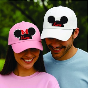 May include: Two baseball caps, one pink and one white. Each cap has a black Mickey Mouse silhouette design with a personalized name in red. The pink cap has a red bow. Both caps are on a person.