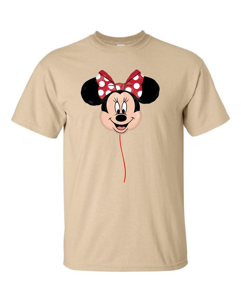 K&ouml;nnte beinhalten: Beiges T-Shirt mit Minnie Mouse-Design. Das Design zeigt Minnie's Gesicht, schwarze Ohren und eine rote Schleife mit wei&szlig;en Punkten. Eine d&uuml;nne rote Linie erstreckt sich vom Gesicht und erzeugt einen Ballon-Effekt.