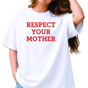 Op de afbeelding: Een wit T-shirt met korte mouwen en de tekst "RESPECT YOUR MOTHER" in dikke rode hoofdletters op de borst gedrukt. Het kledingstuk heeft een ronde hals en een losse pasvorm, gecombineerd met een lichtblauwe spijkerbroek.