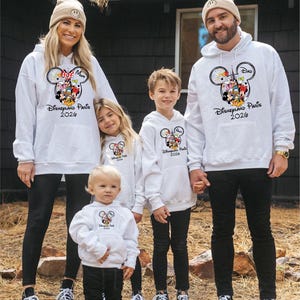 May include: A family of five wearing matching white hoodies with a Mickey Mouse design and the text "Disneyland Paris 2020". The hoodies have a front pocket and are paired with black trousers and trainers. The family is standing in front of a dark building.