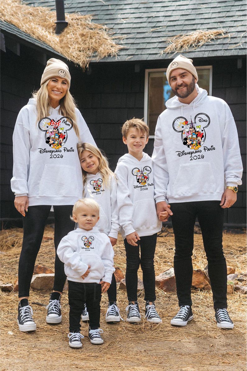 May include: A family of five wearing matching white hoodies with a Mickey Mouse design and the text "Disneyland Paris 2020". The hoodies have a front pocket and are paired with black trousers and trainers. The family is standing in front of a dark building.