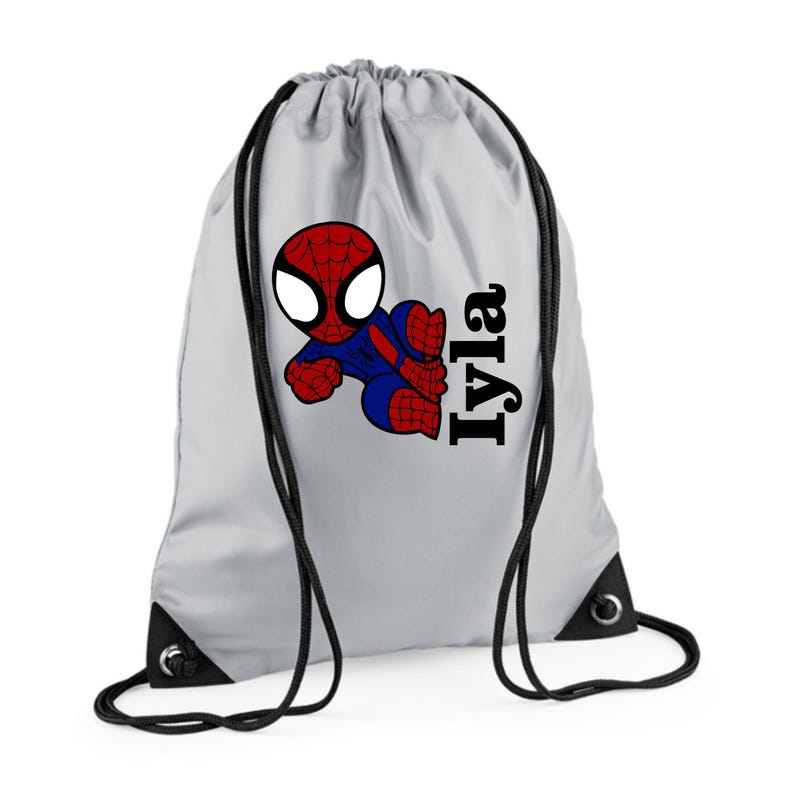 Puede incluir: Una mochila con cord&oacute;n de color gris claro con un dise&ntilde;o de Spider-Man de dibujos animados en rojo, azul y negro. El nombre "Iyla" est&aacute; impreso en negro, con una fuente estilizada en el lado derecho. Cordones y ojales negros completan el dise&ntilde;o.