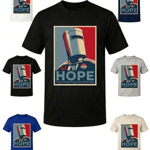 Op de afbeelding: Een collectie t-shirts met korte mouwen in diverse kleuren voorzien van een grafische print van een emmervormige helm. Het ontwerp bevat de tekst HOPE en COUNT BINFACE FOR CLACTON ON SEA, met een rode en blauwe achtergrond achter de helm.