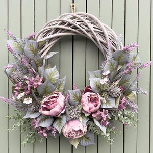 May include: A circular wreath featuring a woven grey base, decorated with pink peonies, lavender and grey-green foliage. The wreath is set against a light green wooden backdrop, offering a natural and welcoming appearance.