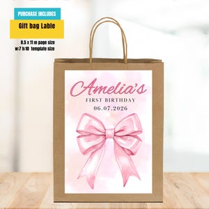 Puede incluir: Una bolsa de regalo de papel marr&oacute;n con un dise&ntilde;o de lazo rosa y el texto "Amelia's FIRST BIRTHDAY 06.07.2026". La bolsa incluye una etiqueta de bolsa de regalo con un tama&ntilde;o de p&aacute;gina de 21,6 x 27,9 cm y un tama&ntilde;o de plantilla de a:17,8 a:25,4 cm.