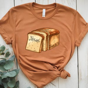May include: A brown t-shirt with a graphic of bread slices. The bread slices have the word "Jesus" written on one slice and the text "John 6:35" on another slice.
