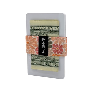 May include: A white card holder with a floral patterned elastic band holding two dollar bills. The elastic band is peach and pink with the text "BANDit!" in black.