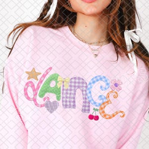 Könnte beinhalten: Hellrosa Sweatshirt mit dem Wort "Dance" in einem Patchwork-Design. Die Buchstaben sind mit Sternen, Herzen, Schleifen und Blumen verziert. Das Sweatshirt hat lange Ärmel und einen Rundhalsausschnitt.