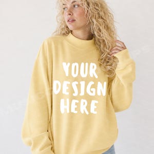 May include: A pale yellow long-sleeve crewneck sweatshirt featuring the white text YOUR DESIGN HERE printed in a bold, rounded sans-serif font across the chest. The garment has a ribbed collar, cuffs, and hem, with a relaxed fit.