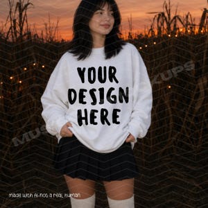Op de afbeelding: Een wit sweatshirt met lange mouwen en een ronde hals, voorzien van de zwarte tekst YOUR DESIGN HERE op de voorzijde. Het sweatshirt wordt gecombineerd met een zwarte geplooide minirok en lichtgekleurde kniekousen voor een casual uitstraling.