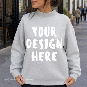 May include: A light gray long-sleeve sweatshirt with a mock neck collar. The front of the garment features the white text "YOUR DESIGN HERE" printed in a bold, sans-serif font. The sweatshirt has long sleeves and a ribbed hem and cuffs.