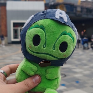 Könnte beinhalten: Ein grünes Plüschfrosch-Spielzeug, das einen Hut im Jeans-Stil mit einem weißen "A" trägt. Der Frosch hat große schwarze Augen und ein kleines Lächeln. Das Spielzeug wird in der Hand einer Person gehalten.