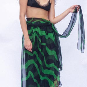 Puede incluir: Un bralette negro con ribete de encaje combinado con una falda transparente a media pantorrilla con un patrón ondulado verde y negro. La falda tiene una cintura estilo envolvente con una larga faja de tela fluida y un dobladillo con volantes, con tacones negros.