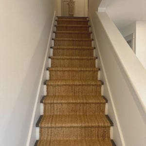 May include: A staircase with woven brown stair runners. The stairs have white risers and dark brown edges. The walls are painted white, and a white banister runs along the right side of the stairs.