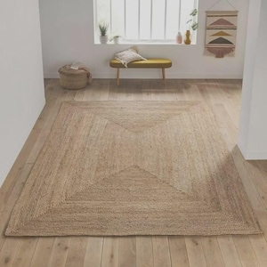 May include: A large, rectangular jute rug with a geometric design. The rug features a square within a square, creating a textured pattern. The natural beige color complements the light wood flooring in the room. The rug is a decorative element for a living space.