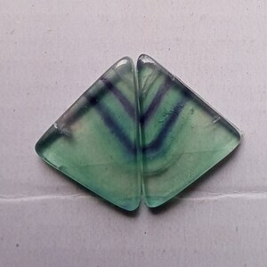 Puede incluir: Dos piezas triangulares de fluorita verde con vetas p&uacute;rpuras y blancas. La fluorita es transl&uacute;cida y tiene un brillo v&iacute;treo.