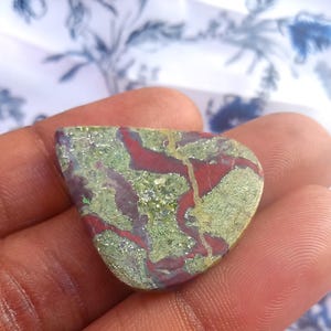 Puede incluir: Un caboch&oacute;n de jaspe verde y rojo con un patr&oacute;n &uacute;nico de colores y vetas en espiral. La piedra es lisa y pulida, con un aspecto natural y org&aacute;nico.