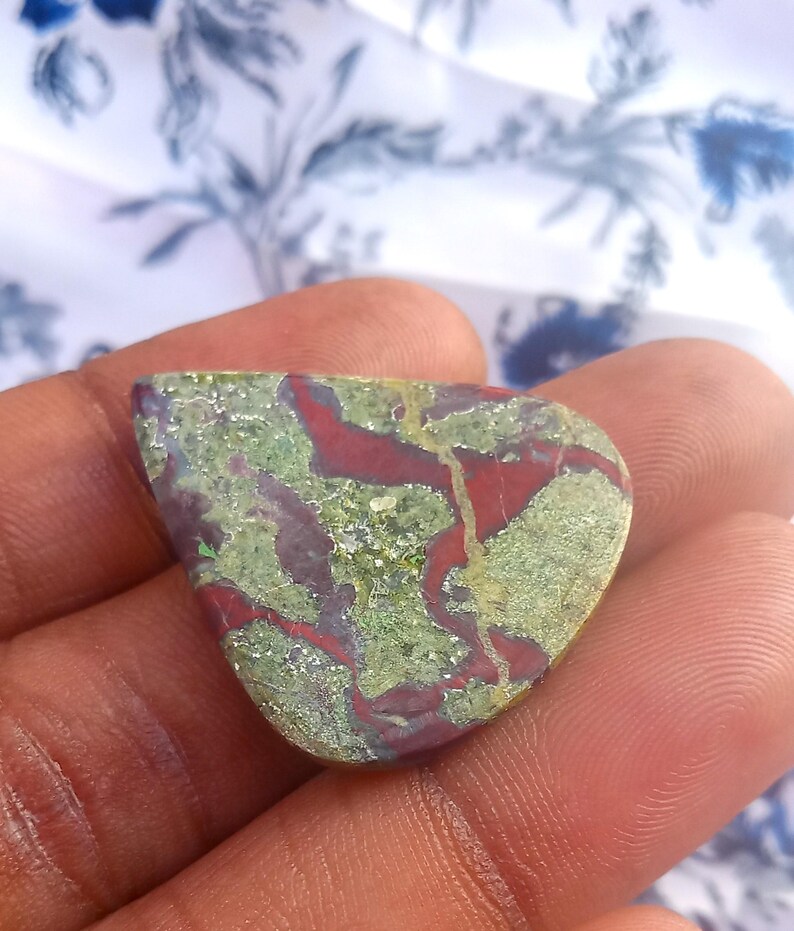Puede incluir: Un caboch&oacute;n de jaspe verde y rojo con un patr&oacute;n &uacute;nico de colores y vetas en espiral. La piedra es lisa y pulida, con un aspecto natural y org&aacute;nico.