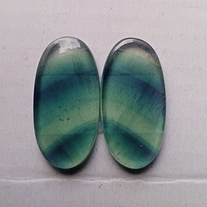 Puede incluir: Dos cabujones ovalados de fluorita verde y azul con un degradado de color verde claro y blanco. Los cabujones son lisos y pulidos.